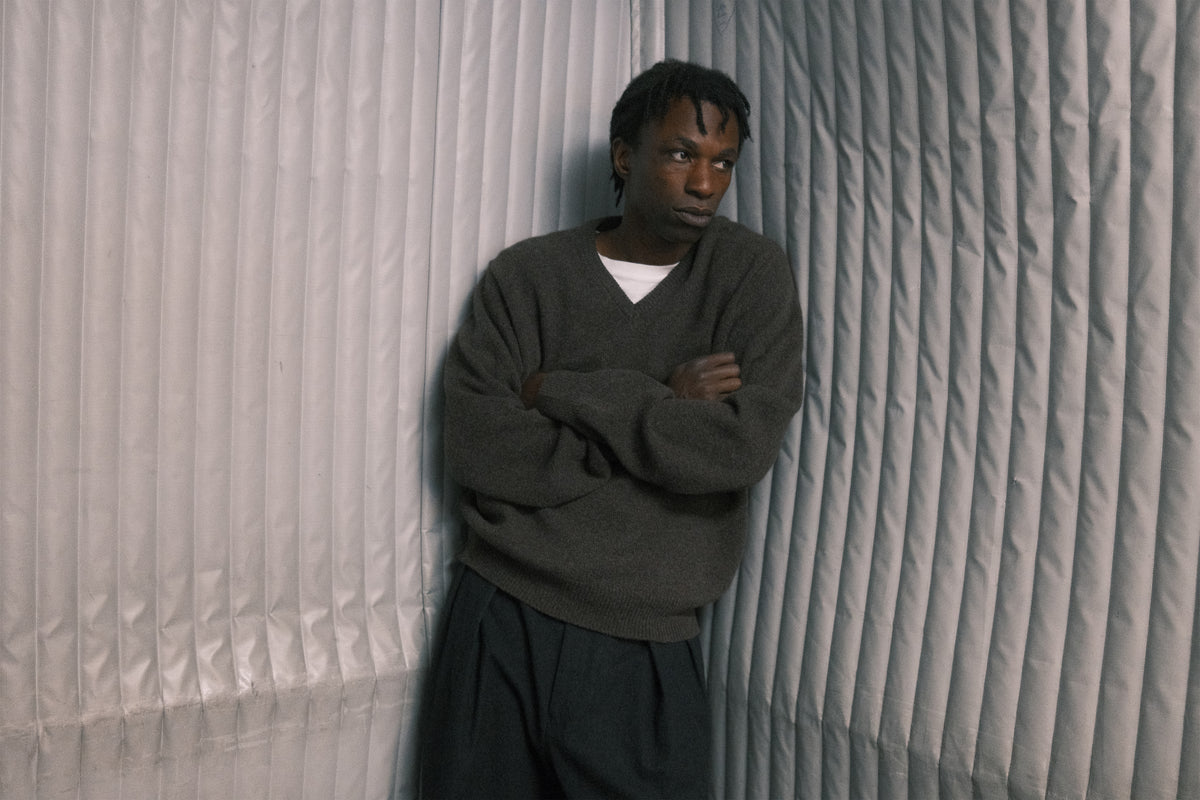 A man stands with arms crossed, wearing a dark gray v-neck sweater over a white undershirt and dark pants, posed against a gray pleated fabric backdrop.