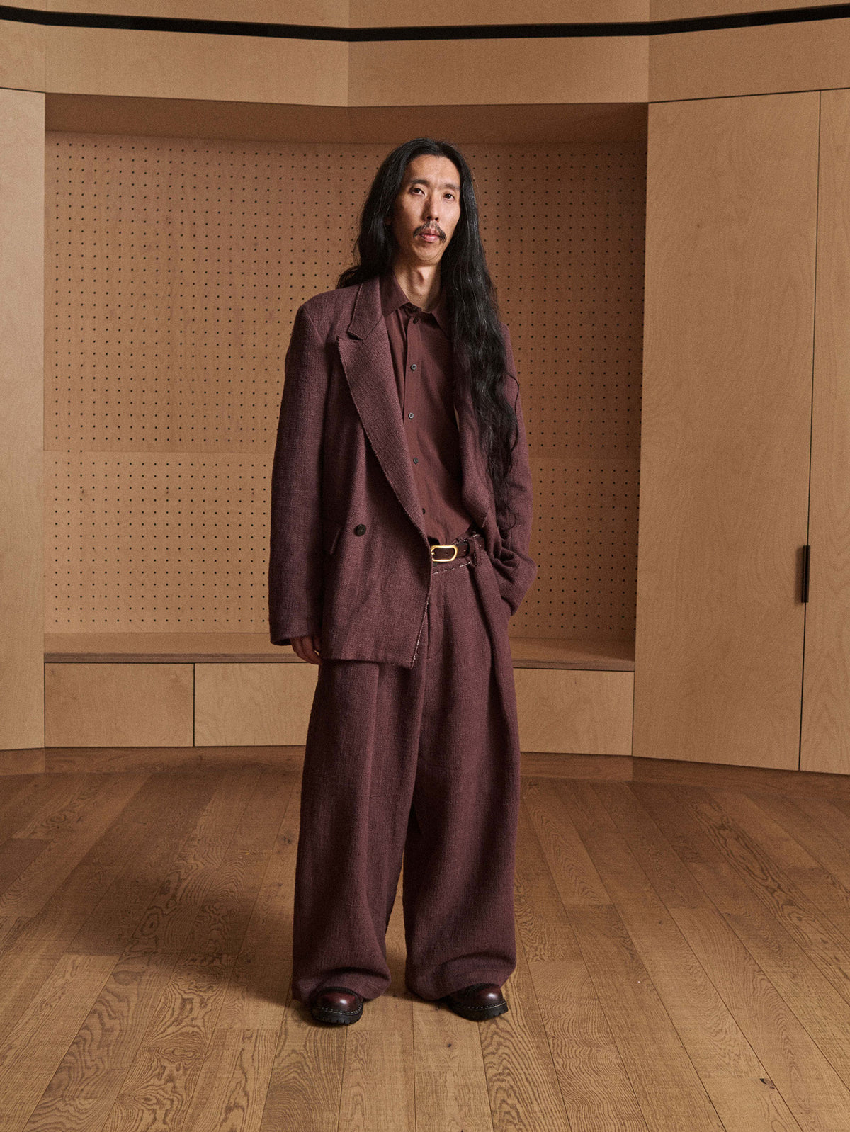 A man with long black hair stands in a minimalist wooden room wearing a matching dark brown linen suit with a double-breasted blazer, collared shirt, and wide-leg trousers.