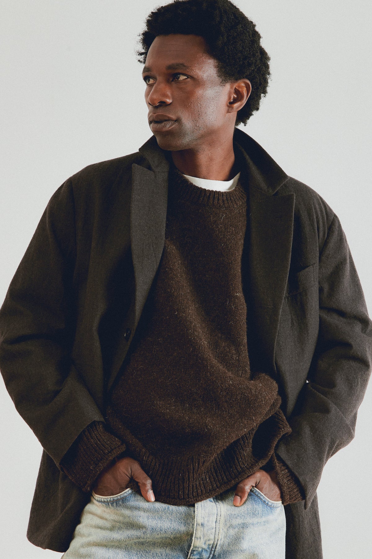 A man wearing a dark brown knit sweater under a dark olive-brown jacket, paired with light-wash blue jeans, photographed against a plain light gray background.