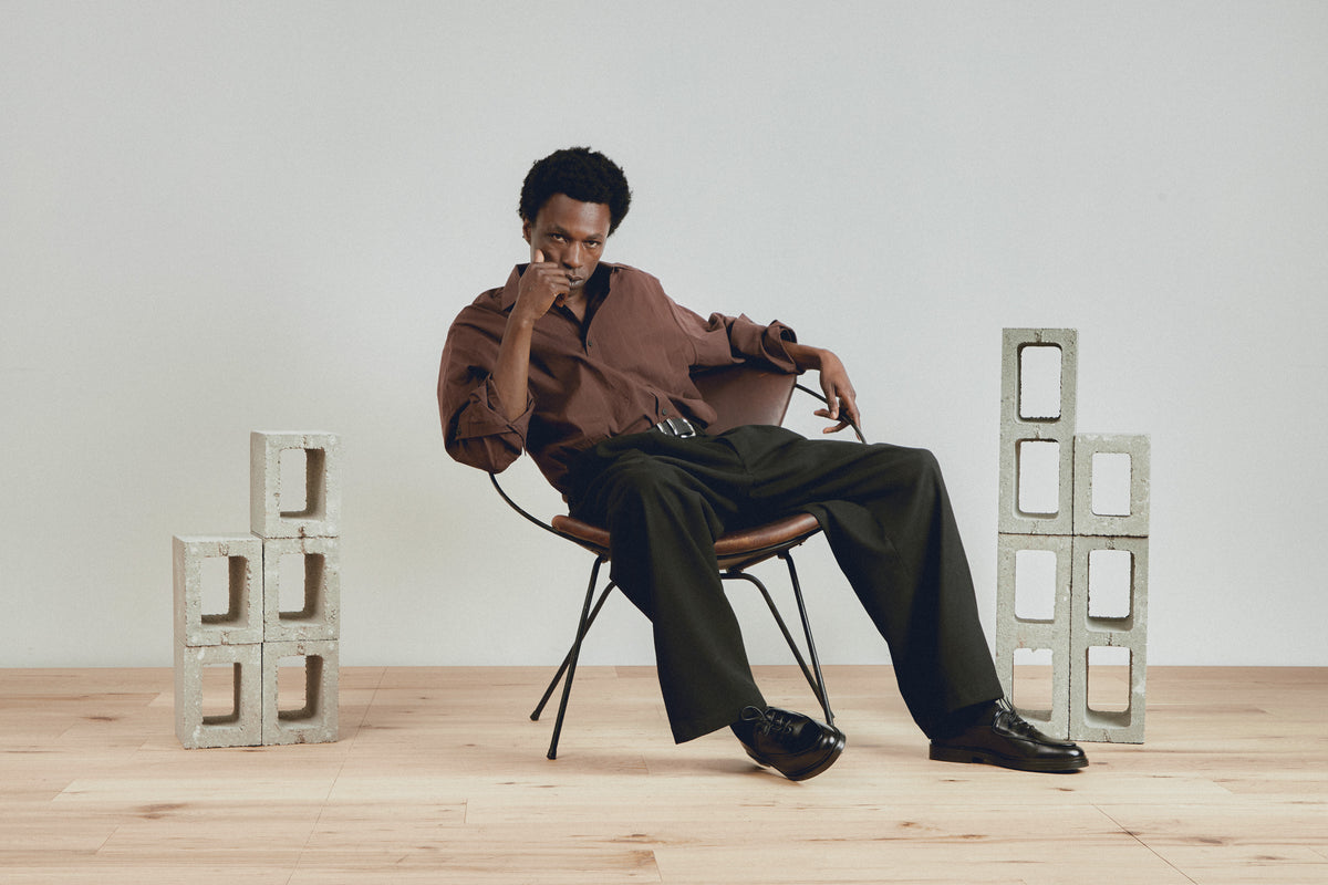 A man sits on a metal chair in a studio setting, wearing a brown button-up shirt and dark trousers. Concrete block props are arranged on either side of him against a light gray wall and wood floor.