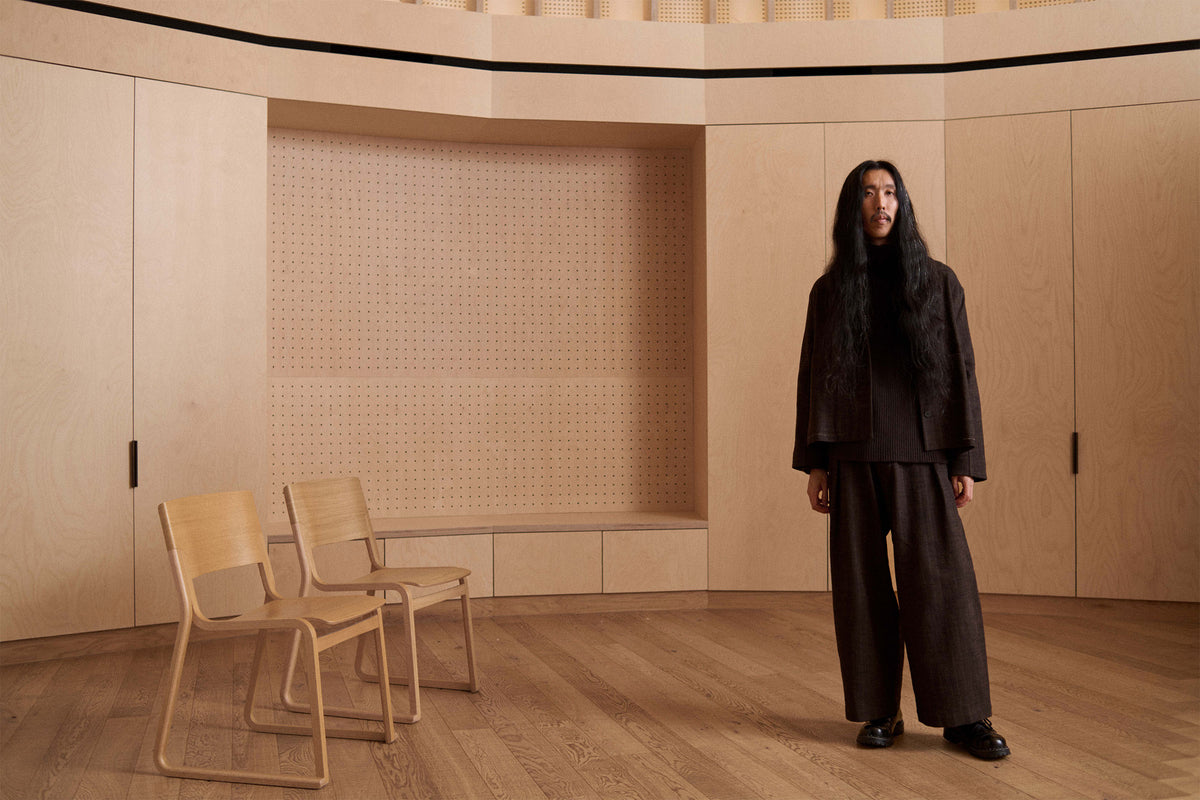 A person with long dark hair stands in a minimalist room with light wood paneling and two wooden chairs, wearing a dark brown or black textured jacket and wide-leg pants.