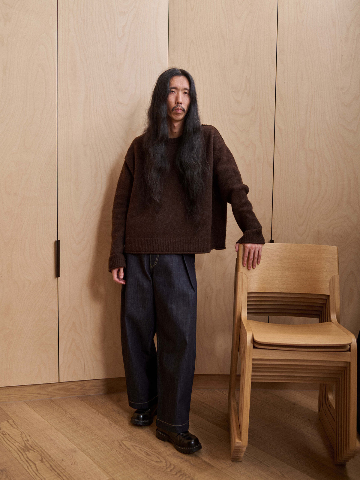 A man with long dark hair stands indoors next to a light wood chair, wearing a dark brown knit sweater and dark blue jeans. The background consists of light wood-paneled walls and a matching wood floor.