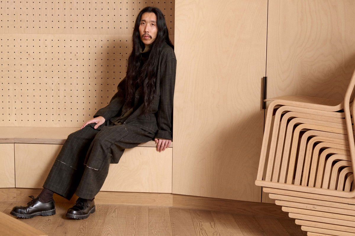 A man with long dark hair sits on a wooden bench in a minimalist interior with light wood paneling. He is wearing a dark oversized cardigan and matching dark trousers, paired with black shoes.